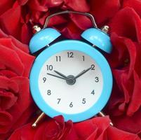 Little cute blue alarm clock surrounded by red roses heads photo