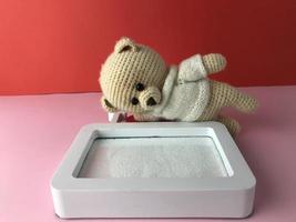 soft knitted bear on a red-pink background with a cute white frame. a bear with black eyes made of beads lies on the background near the photo frame and looks at the photo