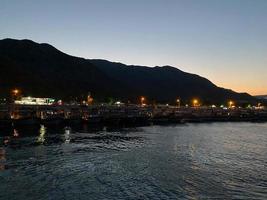 Beautiful coast and beach with outlines of mountains against the backdrop of sunset from the sea or ocean and burning lights of the city and port with ships photo