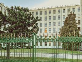 white brick house in the city center. the building is located behind a green metal fence with patterned railings. tall trees grow next to the buildings photo