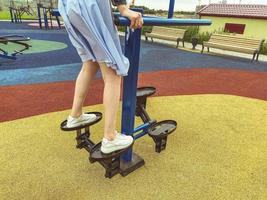 doing sports on the simulator in the park in the fresh air. a girl in a blue dress and white sneakers trains her legs on the platform. active leisure, outdoor time photo