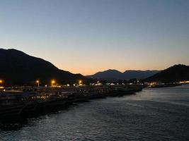 hermosa costa y playa con contornos de montañas contra el telón de fondo de la puesta de sol desde el mar o el océano y luces ardientes de la ciudad y el puerto con barcos foto