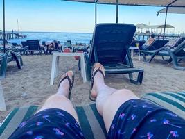 país cálido y tropical con mar y montañas. en la playa hay guijarros y piedras pequeñas. hay una tumbona verde en el suelo, un hombre en pantalones cortos y chanclas descansa sobre ella foto