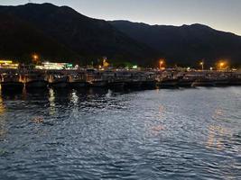 Beautiful coast and beach with outlines of mountains against the backdrop of sunset from the sea or ocean and burning lights of the city and port with ships photo