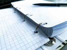 A writing pen rests on a notepad with squared paper sheets on a work desk with stationery in a business office photo