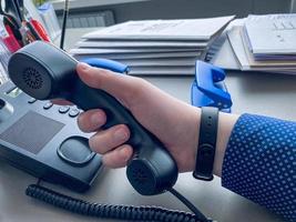 un hombre con una camisa azul con un patrón toma un teléfono negro para hacer una llamada. telefono antiguo. centro de llamadas. trabajo de oficina. teléfono con botones y altavoz foto