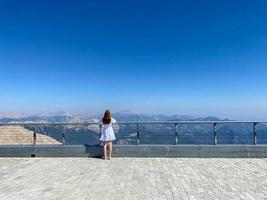 Back view of young Asian female tourist in black top and grey pants looking at camera standing next to fence on observation deck with green hills and blue sky in background photo
