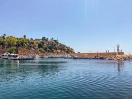 clear, blue water of the sea. an island with a lighthouse in the sea. near a high rock with houses of local residents. yachts and ships are in the port photo