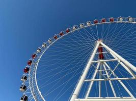 A new very tall white Ferris wheel with comfortable booths for people is slowly rotating in a circle in the center of a big city against a clear blue sky photo