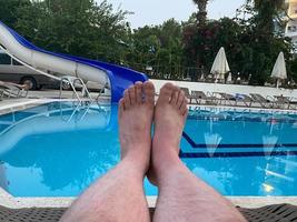 Male tanned legs lie on a beach lounger on a background of a swimming pool with blue water. Summer vacation. Man rest on a sunbed and sunbathes photo