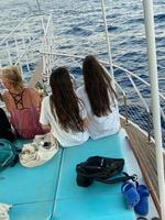 View from back of two beautiful girls standing and looking at camera. Slender and attractive female friends smiling and posing at boat bow. Charming brunettes wearing sunglasses and white clothes photo