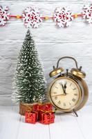 Decorative christmas tree, gift boxes and alarm clock on white wooden background.