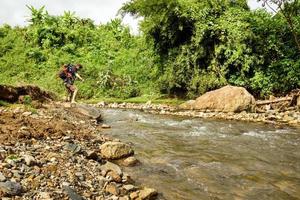 Young white man with backpack crosses the mountain river on a summer sunny day. photo