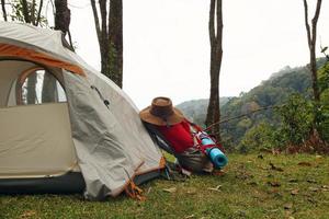 A backpack with a mat and a hat near to a tent on a grass in a camping on a background of mountains and forest. photo