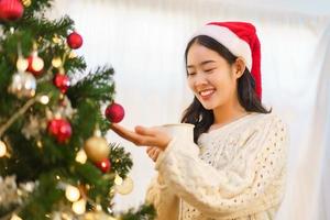 Christmas celebration concept, Asian woman drinks coffee and looking christmas tree with happiness