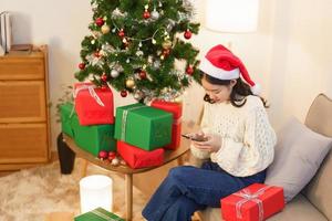 concepto de navidad, una mujer asiática se sienta en una sala de estar decorada con navidad y charla en un teléfono inteligente foto