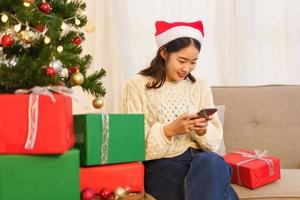 concepto de celebración de navidad, joven asiática sentada en la sala de estar y charlando en el teléfono inteligente foto