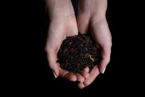 Beautiful female hands hold raw unbrewed tea and offer to try it. Dry tea leaf in hand. Tea with bergamot additives isolated on black photo