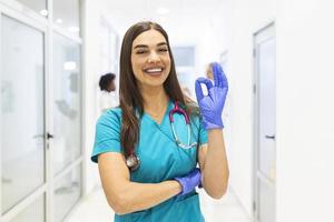 joven doctora en medicina que trabaja en el hospital y en el personal médico, está mostrando un buen signo. la enfermera se para en una pasarela y sonríe con los brazos cruzados. ella está usando matorrales y un estetoscopio. foto