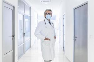 Confident doctor wearing protective face mask while standing in a hallway at the hospital. Senior doctor, surgeon, medic staff professional portrait. photo