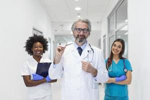 un grupo multiétnico de personal médico. están vestidos con uniformes médicos y batas blancas de laboratorio con estetoscopios alrededor del cuello. algunos de pie en la parte de atrás. el foco está en un hombre maduro en frente foto