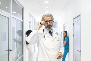 Portrait of confident mature doctor holding his glasses in hospital corridor. He is wearing lab coat in hospital. photo
