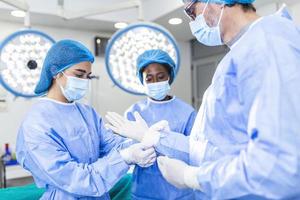 joven enfermera con máscara protectora y ropa de trabajo que ayuda al cirujano con guantes mientras ambos se preparan para la operación