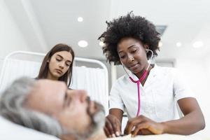 Sick man patient with African American female doctor listens his chest with stethoscope in hospital emergency room. Doctor and nurse come to visit elder patient at hospital room. photo