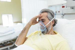 Male coronavirus patient looking away while resting on hospital bed. Man is wearing oxygen mask. He is in hospital during pandemic. photo