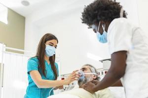 la enfermera pone una máscara de oxígeno en un paciente anciano acostado en la cama de la habitación del hospital mientras un médico afroamericano examina al paciente con estetoscopio, concepto de protección coronavirus covid 19 foto