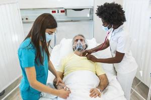 Doctors with protective masks are examining the infected aging patient in the hospital. In the Hospital Senior Patient Rests with oxygen mask, Lying on the Bed. photo