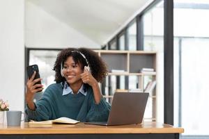 Photo of cheerful joyful mixed-race woman  playing games and wearing wireless headphones at home  Play game on your smartphone that wins the moment of victory. Fun and enjoyable vacation