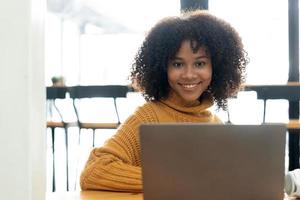 Photo of cheerful joyful mixed-race woman in yellow shirt smiling work on laptop talk speak video  call online. Smart ethnic female in earphones study distant on computer at home. Education concept.