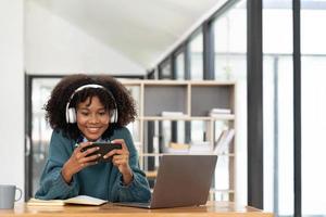 Photo of cheerful joyful mixed-race woman  playing games and wearing wireless headphones at home  Play game on your smartphone that wins the moment of victory. Fun and enjoyable vacation