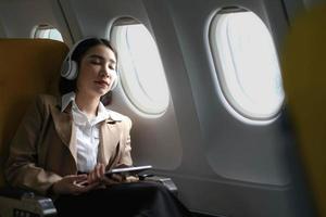 Young caucasian smiling female enjoying her comfortable flight while sitting in airplane cabin, listening to music in earphones and drinking water. Wifi internet access on board, passenger near window