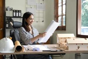Architecture, building, construction and real estate concept - Happy smiling female architect with blueprint, ruler and living house model on table working at home office. Architect woman. photo