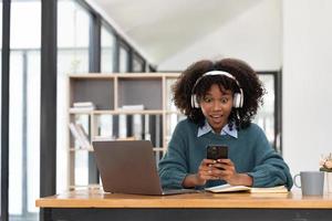 Photo of cheerful joyful mixed-race woman  playing games and wearing wireless headphones at home  Play game on your smartphone that wins the moment of victory. Fun and enjoyable vacation