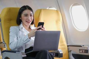 Asian woman passenger sitting in airplane near window and reading news from social networks or using travel applications in smartphone photo