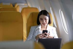 Asian woman passenger sitting in airplane near window and reading news from social networks or using travel applications in smartphone photo