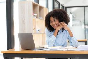 sonriente joven empresaria africana sentada en un escritorio en su oficina trabajando en línea con una laptop foto