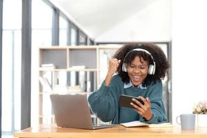 Photo of cheerful joyful mixed-race woman  playing games and wearing wireless headphones at home  Play game on your smartphone that wins the moment of victory. Fun and enjoyable vacation