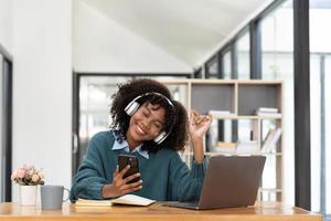 Photo of cheerful joyful mixed-race woman  playing games and wearing wireless headphones at home  Play game on your smartphone that wins the moment of victory. Fun and enjoyable vacation