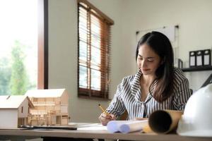 Architecture, building, construction and real estate concept - Happy smiling female architect with blueprint, ruler and living house model on table working at home office. Architect woman. photo