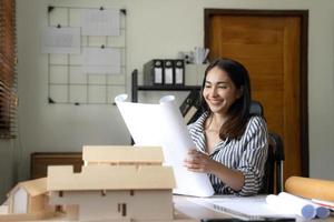 Architecture, building, construction and real estate concept - Happy smiling female architect with blueprint, ruler and living house model on table working at home office. Architect woman. photo