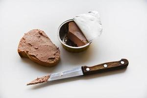 Meat pate in a jar on a white background. Spreading pate on bread with a knife. Meat pate, knife and bread. Delicious breakfast of pate. photo