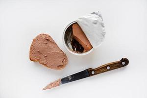 Meat pate in a jar on a white background. Spreading pate on bread with a knife. Meat pate, knife and bread. Delicious breakfast of pate. photo