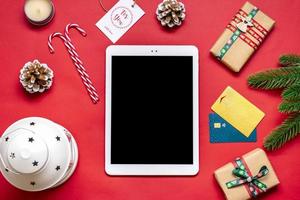 Tablet with black screen, debit card, Xmas decor, bag, boxes on red table Top view Flat lay Holiday shopping list, Happy New Year, Christmas online shop, chooses gifts, makes purchases concept Mock up photo