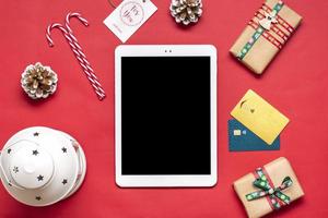 Tablet with black screen, debit card, Xmas decor, bag, boxes on red table Top view Flat lay Holiday shopping list, Happy New Year, Christmas online shop, chooses gifts, makes purchases concept Mock up photo