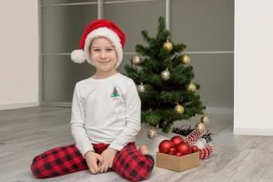 niña en pijama a cuadros y un sombrero de santa se sienta al lado del árbol de navidad y sonríe, navidad foto