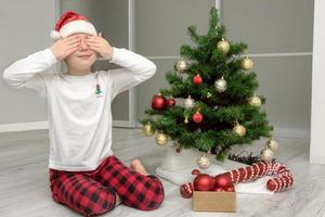 girl in pajamas and a santa hat sits by the christmas tree with her eyes closed, christmas content photo